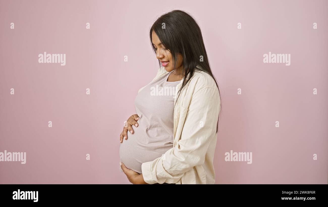 Fröhliche junge schwangere Frau mit einem strahlenden Lächeln berührt freudig ihren Bauch und steht beiläufig über rosa isoliertem Hintergrund Stockfoto
