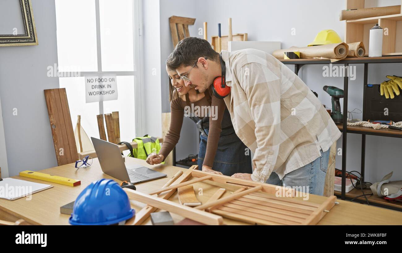 Ein Mann und eine Frau arbeiten in einer Holzwerkstatt zusammen und untersuchen intensiv einen Holzrahmen in der Nähe verschiedener Zimmermannswerkzeuge und Sicherheitsausrüstungen. Stockfoto