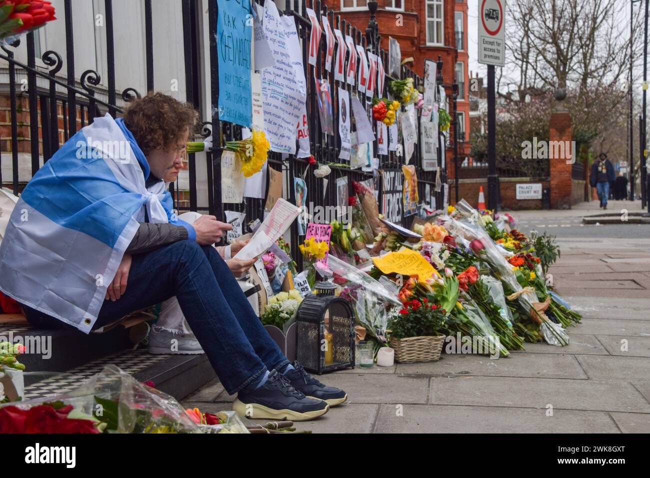 London, England, Großbritannien. Februar 2024. Ein Mann mit einer symbolischen russischen Antikriegsfahne sitzt an der provisorischen Gedenkstätte, während Anhänger von Alexej Nawalny weiterhin Blumen und Tribut gegenüber der russischen Botschaft in London hinterlassen, nachdem der Oppositionsführer im Gefängnis in Russland gestorben war. Quelle: ZUMA Press, Inc./Alamy Live News Stockfoto