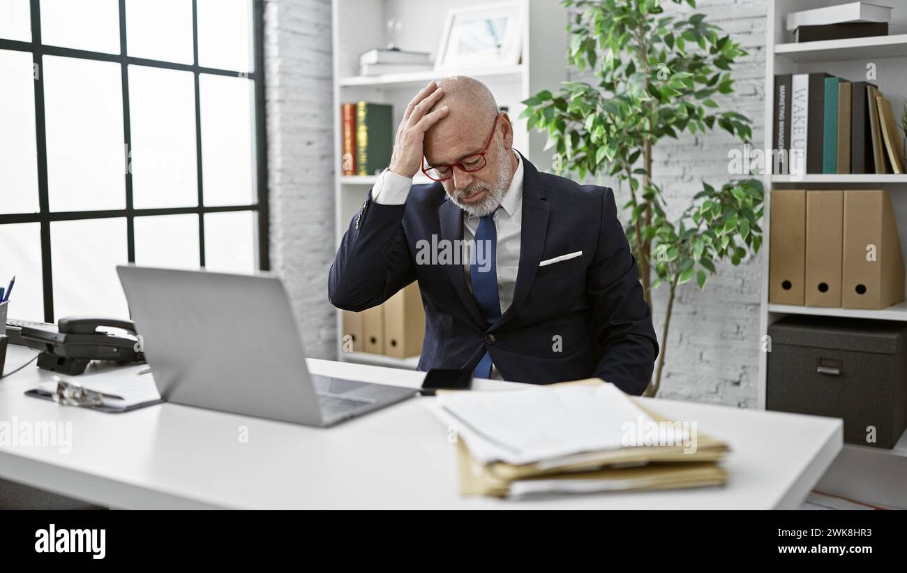 Mann im Anzug mit Glatze, gestresst am Schreibtisch mit Laptop in heller Büroumgebung Stockfoto