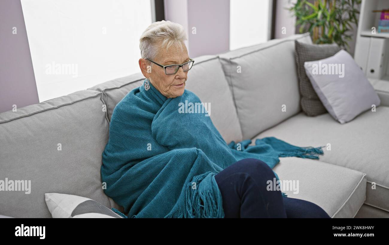 Ältere grauhaarige Frau, die Wärme sucht, in eine Decke gekuschelt bei eisiger Temperatur drinnen, mit Anzeichen von Grippe in ihrem gemütlichen Zuhause Stockfoto