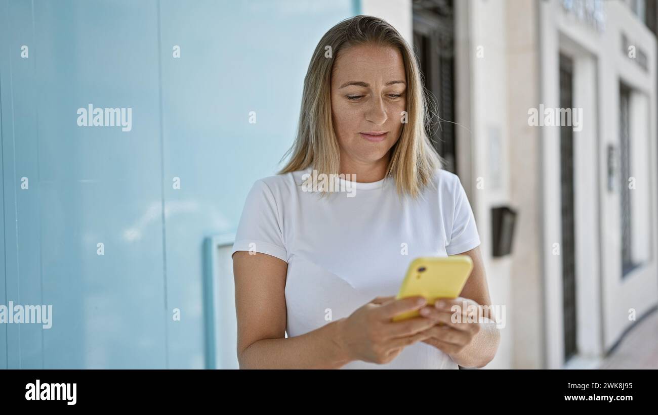 Stilvolle, schöne junge blonde Frau, die sich ernsthaft darauf konzentrierte, Online-Nachrichten auf ihrem Smartphone zu schreiben, stand draußen auf der sonnigen Stadtstraße und lebte Stockfoto