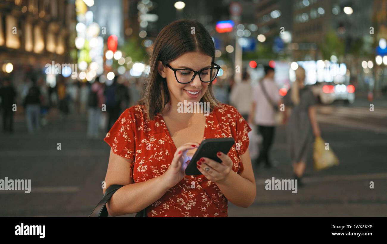 Fröhliche und schöne hispanische Frau, in einem fröhlichen Moment festgehalten, auf ihrem Smartphone unter den Nachtlichtern der städtischen osaka Street SMS geschrieben und verkörpert Stockfoto