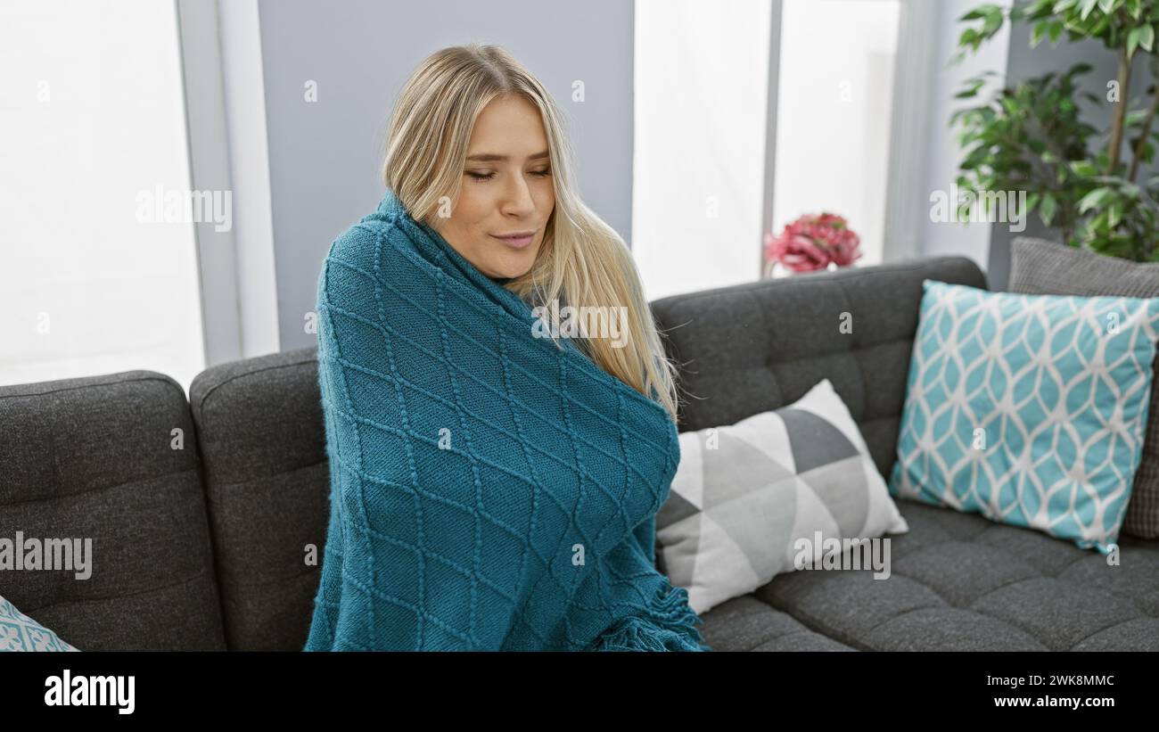 Schöne junge blonde Frau, die in der gemütlichen Wärme ihrer Wohnung friert, auf dem Sofa sitzt, in einer Decke gewickelt, die gegen das Erkältungsfieber der Grippe kämpft Stockfoto