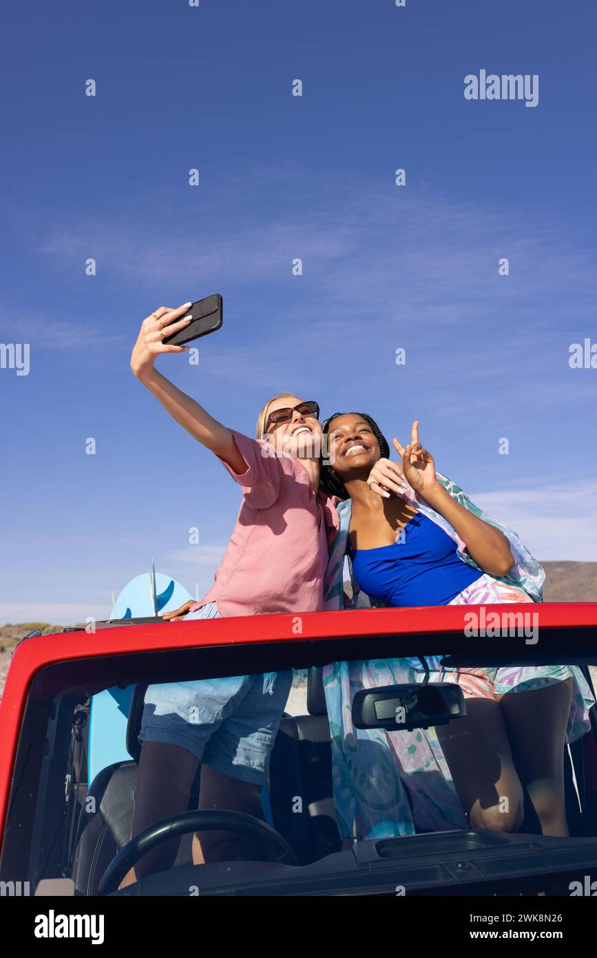 Zwei Frauen verschiedener Rassen machen ein fröhliches Selfie auf einem Roadtrip unter blauem Himmel. Stockfoto