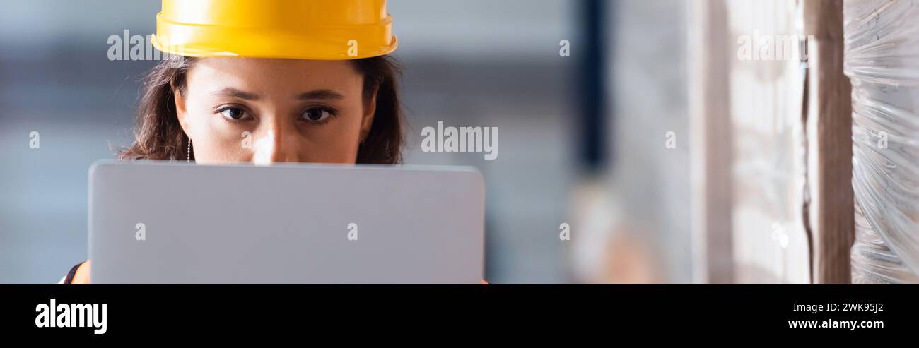 Fokussierte Logistikarbeiterin mit Laptop und Blick auf Kamera, Lagerkontrolle und Frachtüberprüfungsbanner Stockfoto