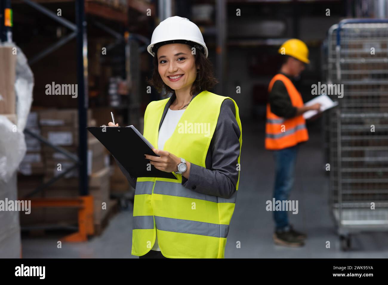 Glücklicher Lagerarbeiter in Sicherheitsweste und mit Schutzhelm auf Klemmbrett, Logistik und Distribution Stockfoto