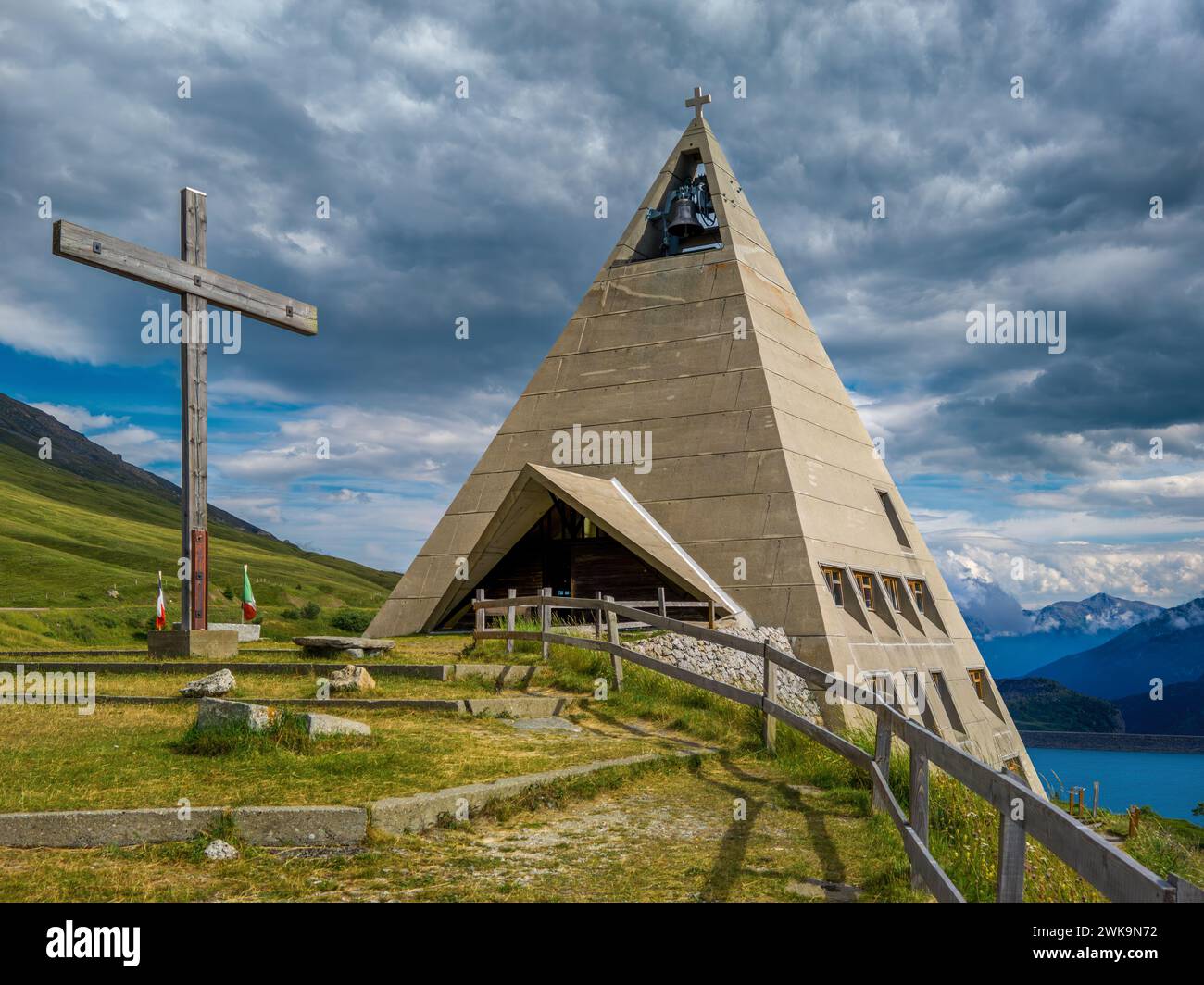 Moderne pyramidenförmige Kirche und Holzkreuz als alpiner See Mont-Cenis und Berge auf dem Hintergrund in Frankreich. Stockfoto