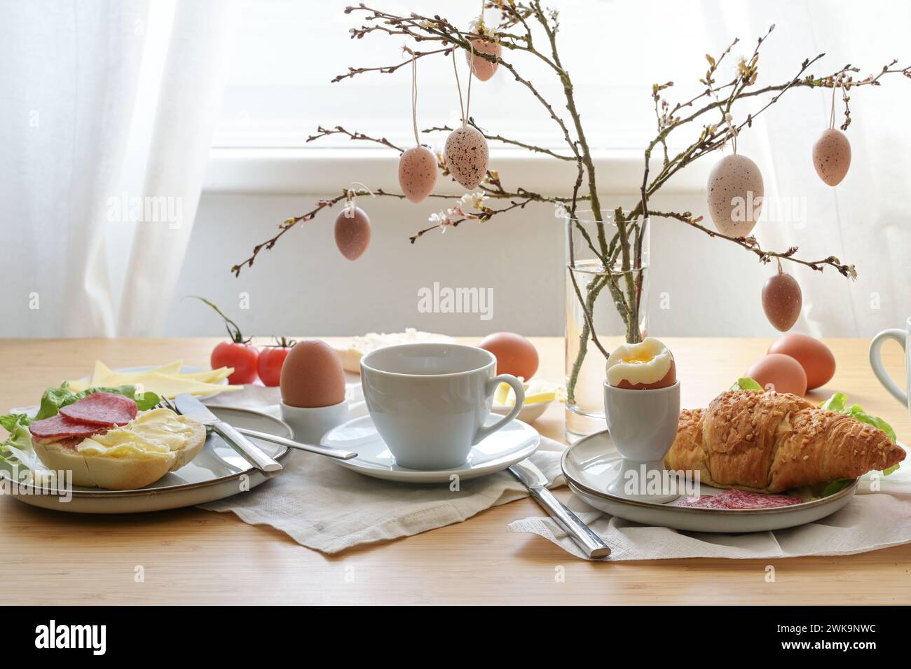 Osterfrühstück mit Ei, Brötchen, Kaffee, Croissant, Käse und Wurst, dekoriert mit Frühlingszweigen und hängenden ostereiern in einer Glasvase auf einem Tab Stockfoto
