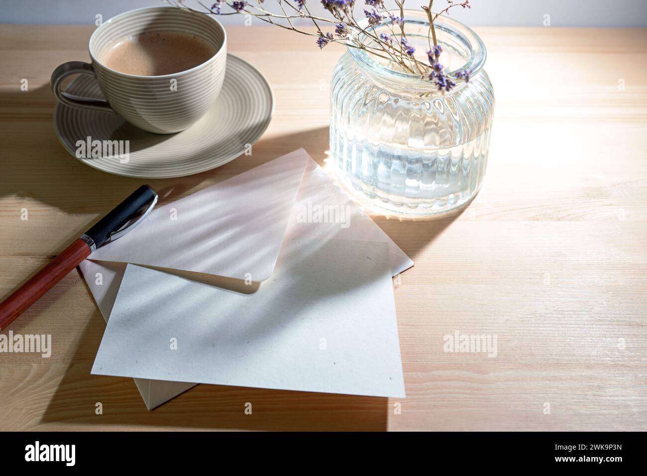Grußkarte und Umschlag aus natürlichem Recyclingpapier, Stift, Kaffeetasse und eine Glasvase mit Sonnenlichtreflexionen am Morgen auf einem Holztisch, W Stockfoto