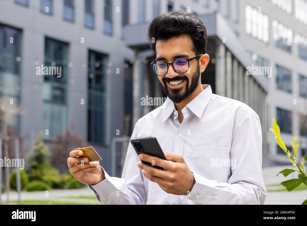 Ein junger Mann mit einem Telefon und einer Bankkreditkarte in den Händen, ein glücklicher Einkäufer wählt Produkte in einem Online-Shop aus, sitzt vor einem Bürogebäude, ein Geschäftsmann läuft durch die Stadt. Stockfoto