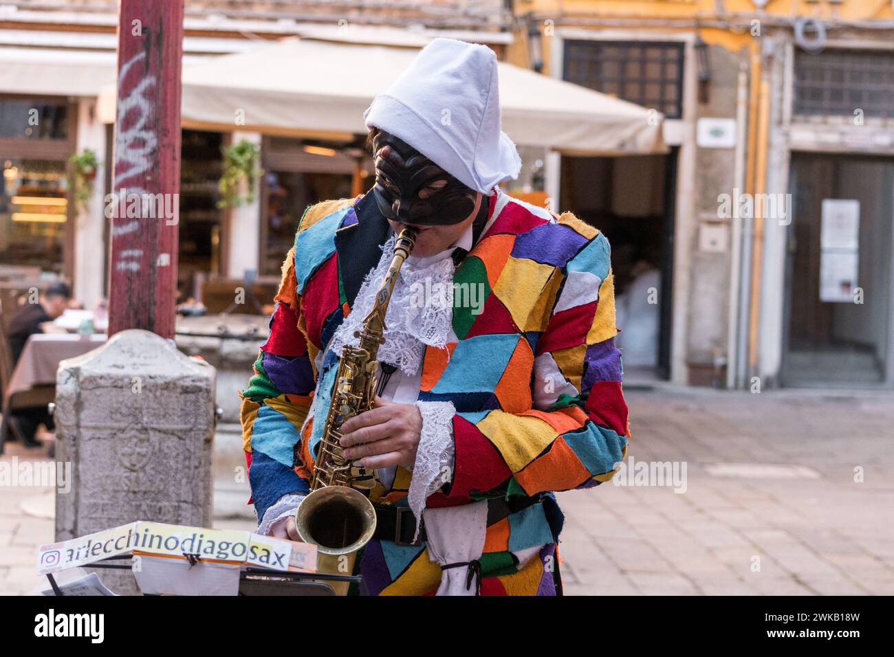 Sonniger Tag am Karneval von Venedig mit wunderschönen Masken und einem Straßenkünstler, der als Harlekin verkleidet ist und Saxophon spielt. ArlecchinoDiagoSax. Stockfoto