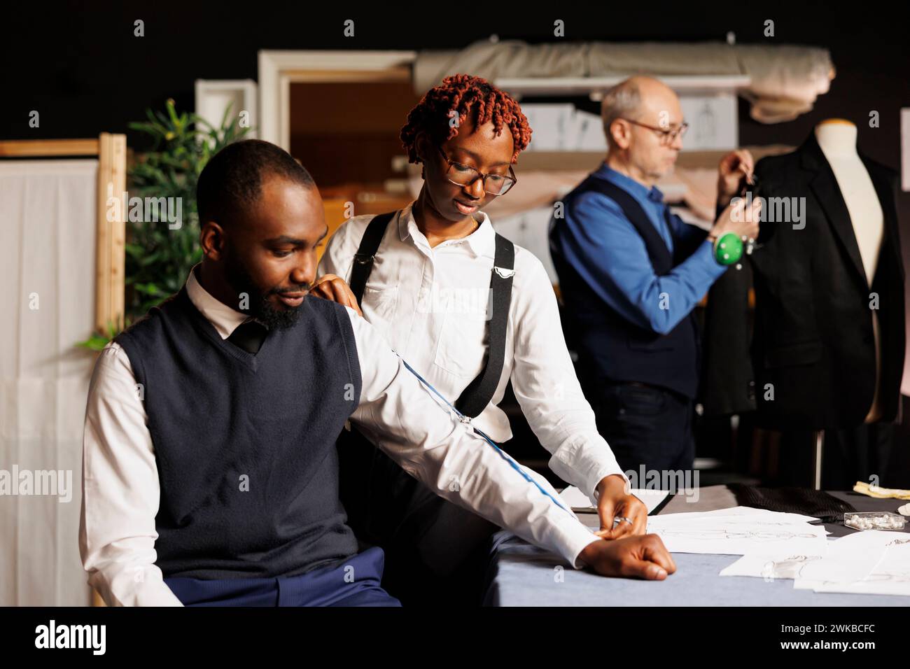 Suitmaker nimmt Kundenmessungen für maßgeschneiderte, handgefertigte Business-Anzüge vor, während ältere Handwerker Anpassungen an der Kleidung vornehmen. Teamwork schneidet hochwertige Kleidung her Stockfoto