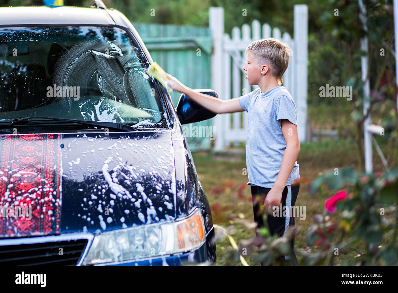 Kaukasischer Teenager wäscht Auto, indem er es mit Waschmittel im Hof des Hauses an einem Sommertag seift. Kinderhilfe Stockfoto
