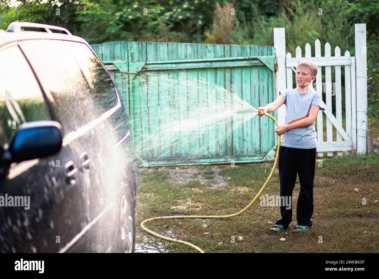 Ein Teenager wäscht ein Auto, indem er Wasser mit Wasser aus einem Gartenschlauch darauf gießt. Das Konzept der Familienzusammenarbeit Stockfoto