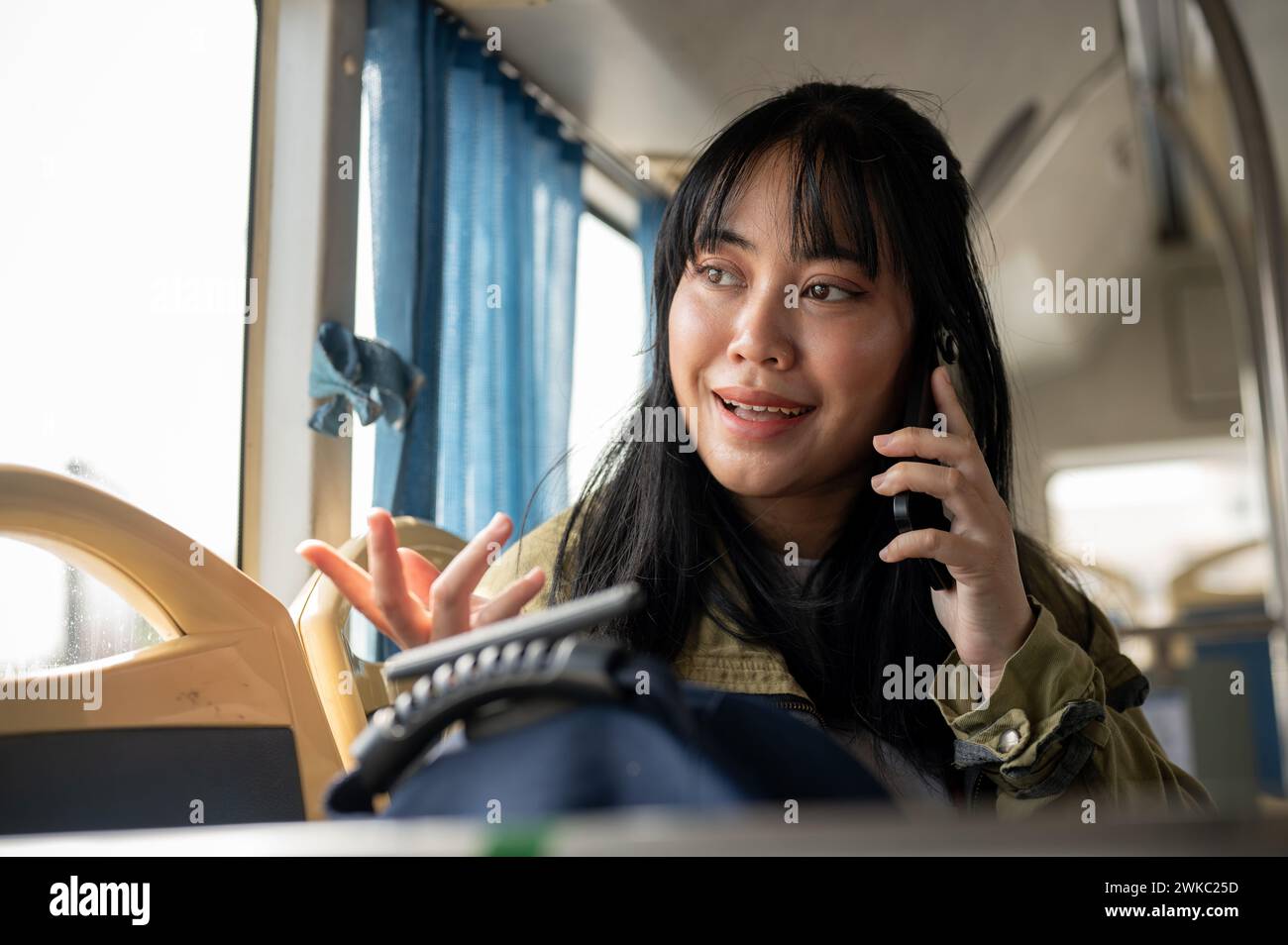 Eine glückliche asiatische Frau sitzt im Bus, blickt aus dem Fenster und bewundert die Aussicht entlang der Straße, während sie telefoniert. Menschen und öffentliche Verkehrsmittel Stockfoto