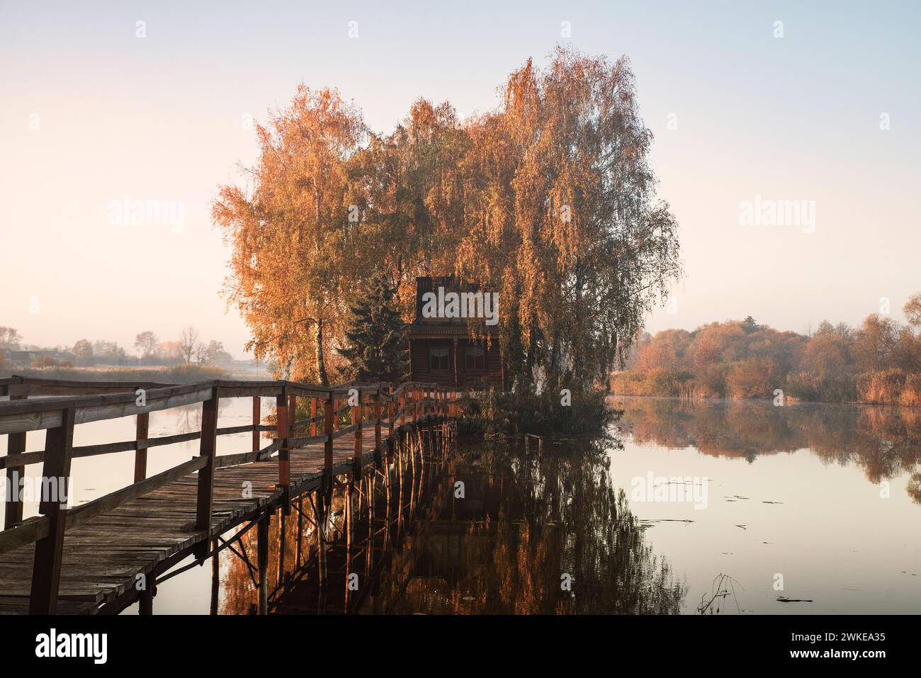 Morgenlandschaft eines kleinen Hauses auf einer künstlichen Insel mit einer langen Holzbrücke. Um das Haus herum ist ein kleiner See mit leichtem Nebel. Herbst ca. Stockfoto