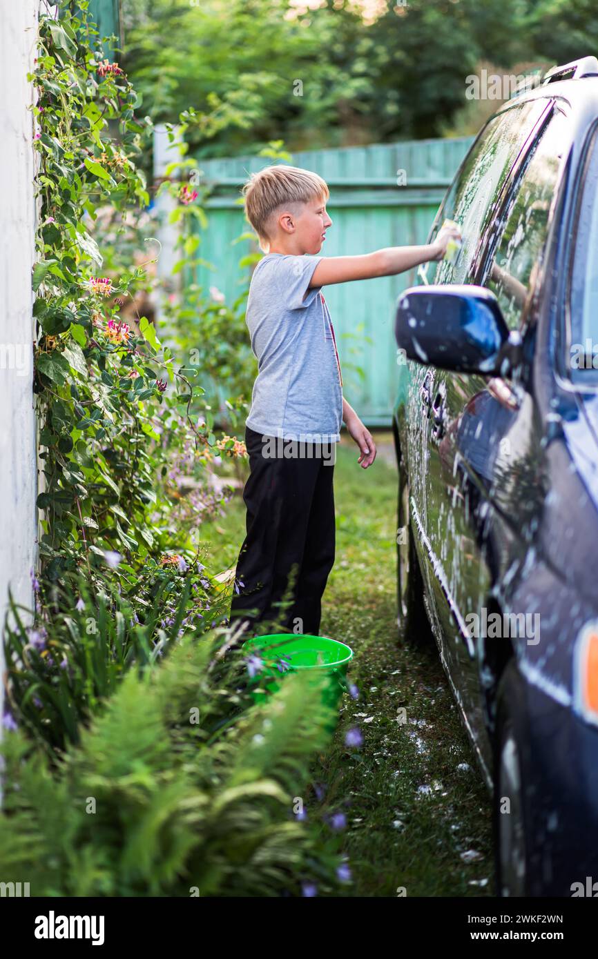 Ein Teenager im Trainingsanzug wäscht im Sommer einen Minivan mit Seifenwasser im Hof eines Hauses. Das Konzept der Förderung von Verantwortung im Alltag Stockfoto