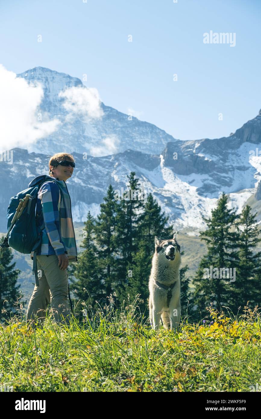 Sportliche Frau mit Huskyhund, die vor den schneebedeckten Bergen steht und den Blick auf die Natur der Schweiz genießt. Fernweh, Sport, Schönheit in der Natur Stockfoto