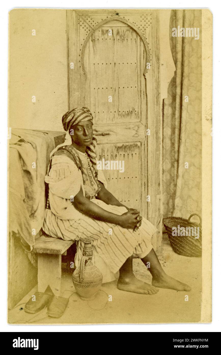 Original viktorianische Epoche CDV Carte de Visite (Visitenkarte oder CDV) Albumenfoto einer schönen jungen Dienerin in traditionellem marokkanischem Kleid, die auf einer Bank mit verzierten Holztüren sitzt, sieht aus wie ein Studio, Tanger, Marokko. 1870er Jahre Stockfoto