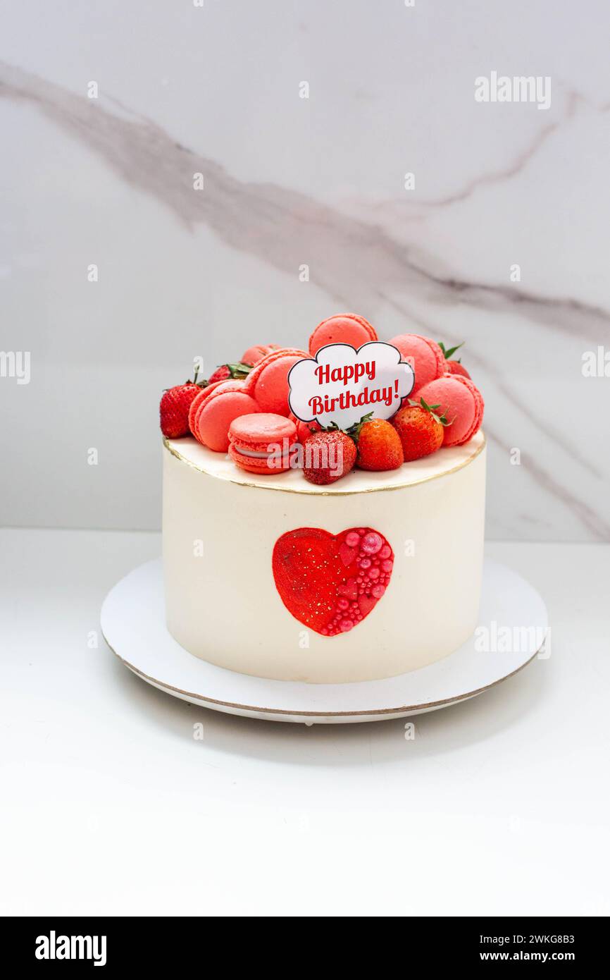 Schöner weißer Kuchen mit rotem Herz, Macarons, frischer Erdbeere und Text auf einfachem Marmorhintergrund. Idee zum romantischen Jubiläum Stockfoto