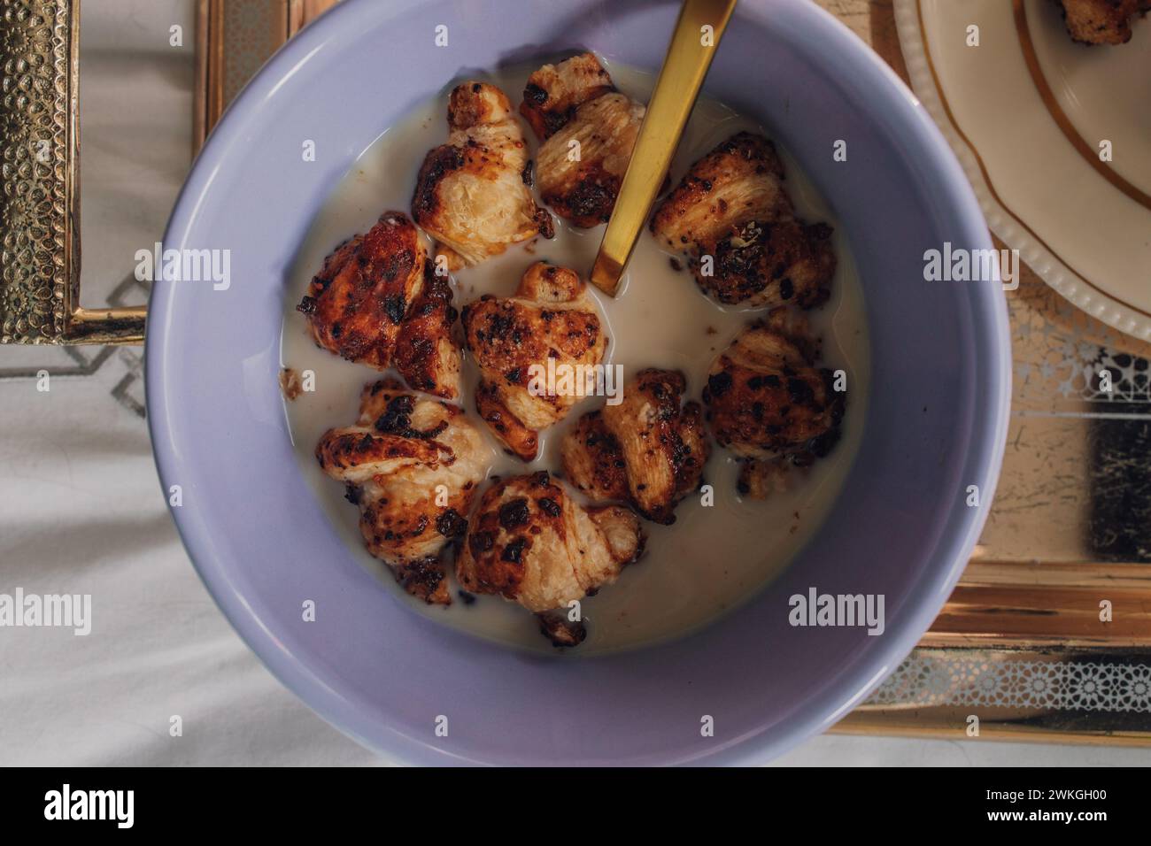 Blick von oben auf die lila Schüssel mit Mini Croissant Müsli und goldenem Löffel auf silbernem Tablett auf dem Bett Stockfoto