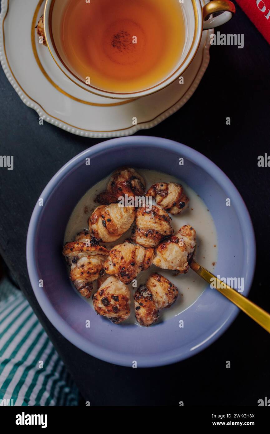 Blick von oben auf die lila Schüssel mit Mini Blaubeeren Zucker Croissant Müsli, goldenem Löffel und weißer Teetasse schwarzen Tees auf schwarzem Tisch Stockfoto