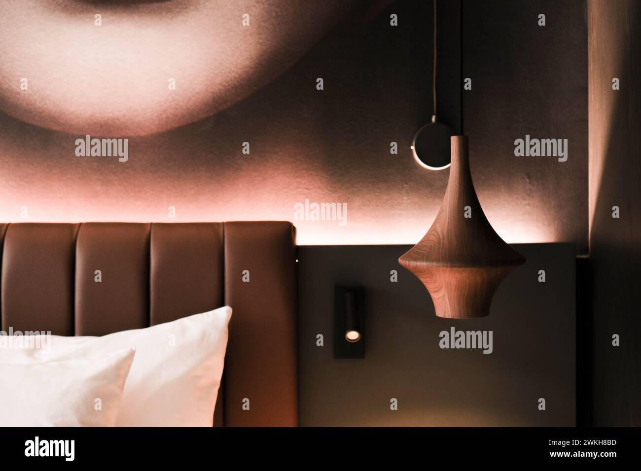 Dieses Bild zeigt einen Teil eines modernen Hotelzimmers mit einem weichen Kopfteil, einer eleganten Pendelleuchte und einer künstlerischen Wanddekoration, die einen zusätzlichen Glanz verleiht Stockfoto