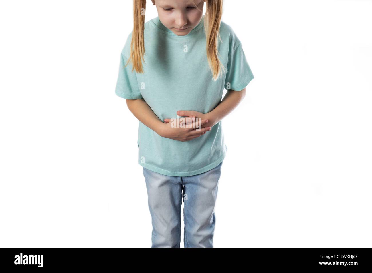 Ein siebenjähriges blondes Mädchen steht und hält ihren Bauch auf weißem Hintergrund. Das Konzept der Bauchschmerzen bei Kindern aufgrund schlechter Ernährung. Infl Stockfoto