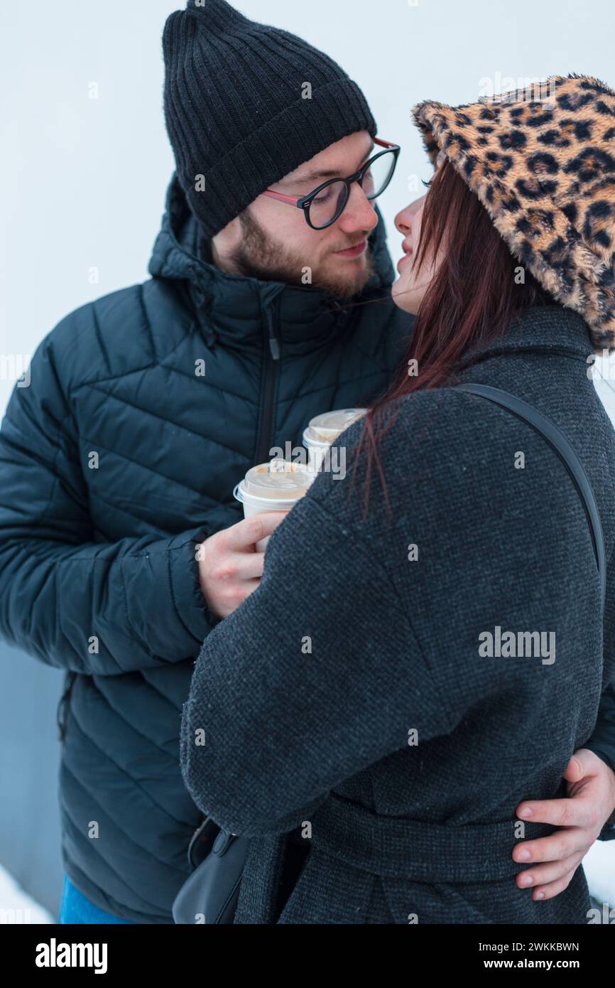 Stilvolle junge Liebhaber von modischer Winterkleidung mit Jacke halten eine Tasse Kaffee und umarmen sich auf weißem Hintergrund Stockfoto