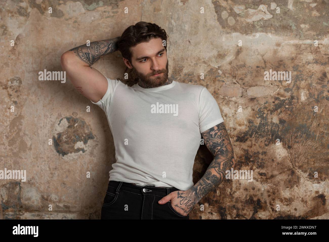 Hübscher muskulöser, brutaler Hipster-Mann mit Frisur und Bart mit einem Tattoo in einem weißen T-Shirt steht neben einer alten Grunge-Betonwand Stockfoto