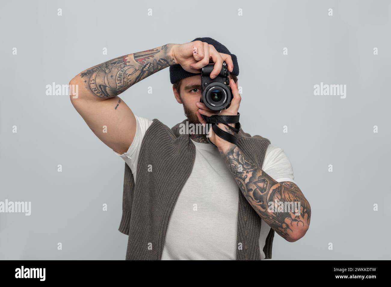 Gutaussehender Mann mit Tätowierung mit Strickmütze mit Strickpullover, der spiegellose Fotokamera hält und Fotos im Studio macht Stockfoto