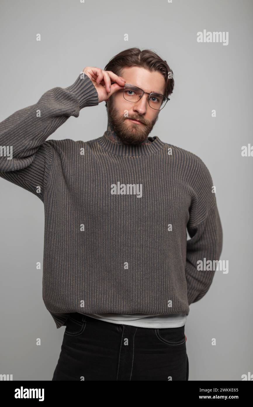 Cooler, hübscher, modischer Hipster-Mann mit Frisur und Bart mit Vintage-Brille in einem modischen Strickpullover passt seine Brille auf eine weiße Ba an Stockfoto
