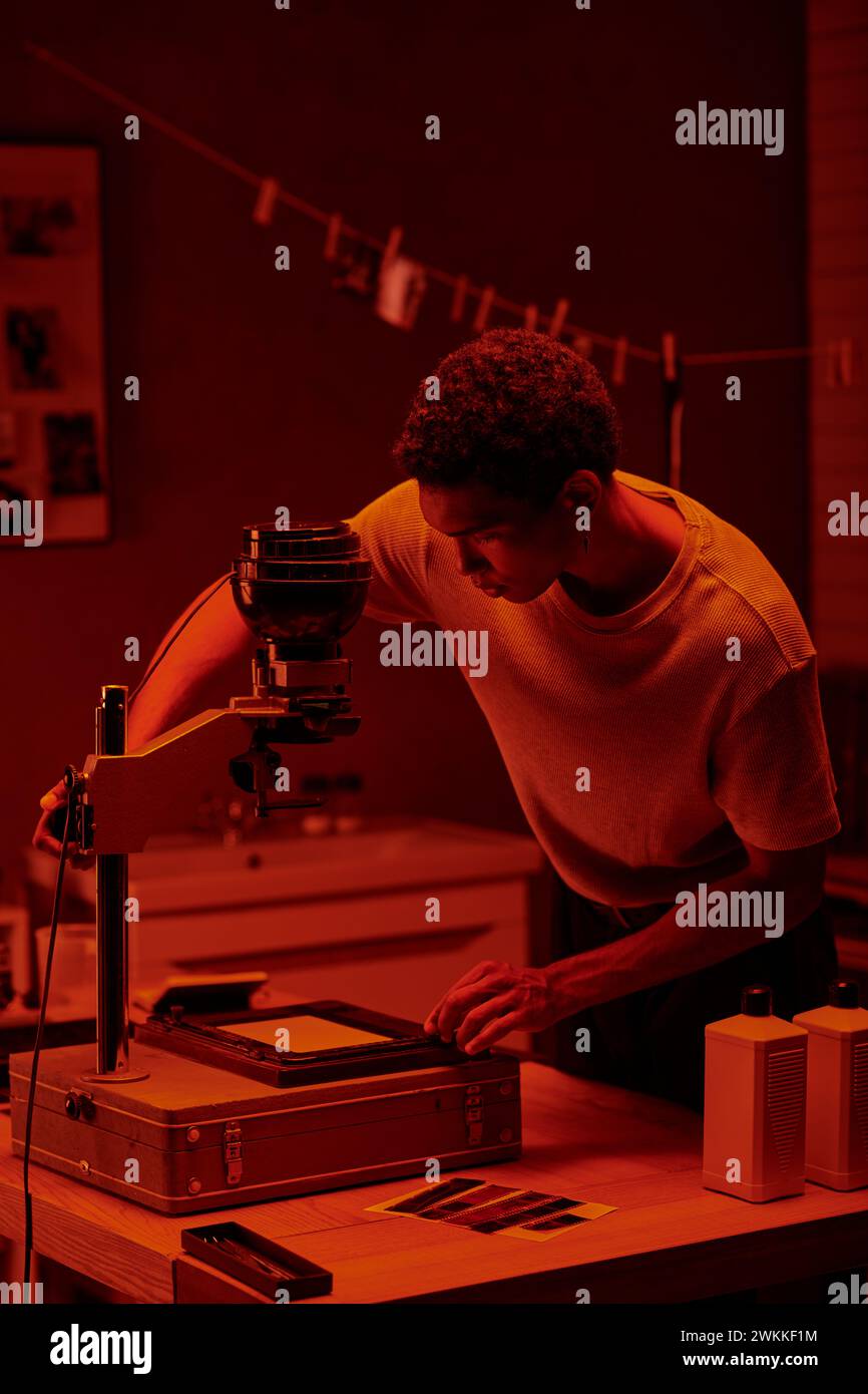 afroamerikanischer Fotograf, der Fotonegativ unter dem roten Sicherheitslicht einer Dunkelkammer inspiziert Stockfoto