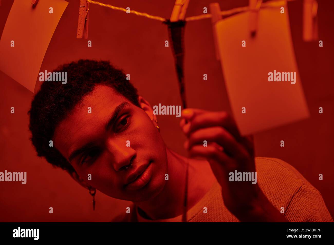 Junger afroamerikanischer Mann, der sich einen frisch entwickelten Filmstreifen in einer rot beleuchteten Dunkelkammer anschaut, Nostalgie Stockfoto