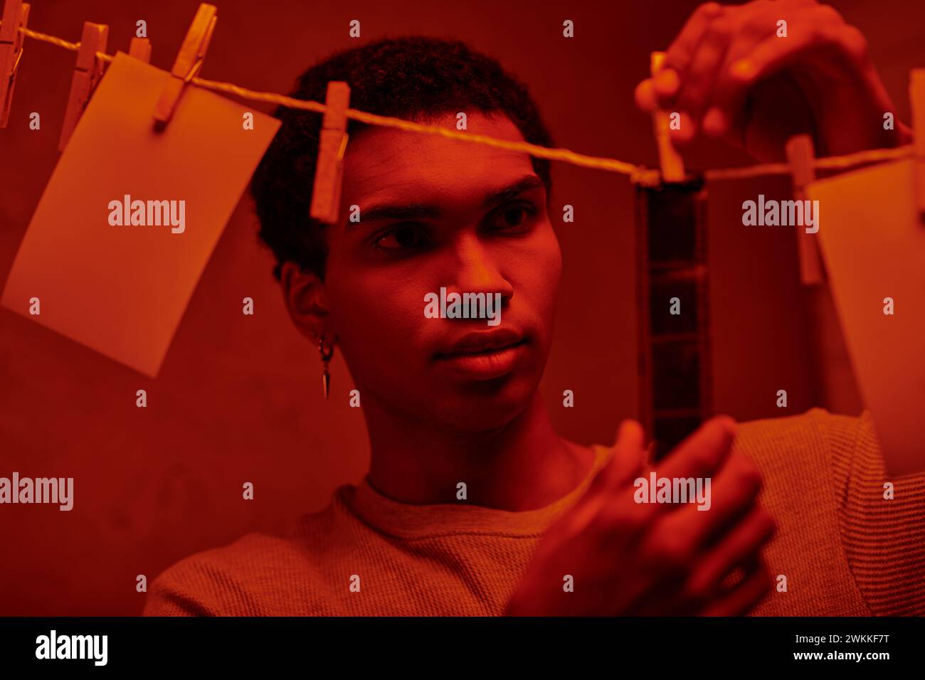 Ein junger afroamerikaner hängt einen frisch entwickelten Filmstreifen in einer rotbeleuchteten Dunkelkammer, zeitlos Stockfoto