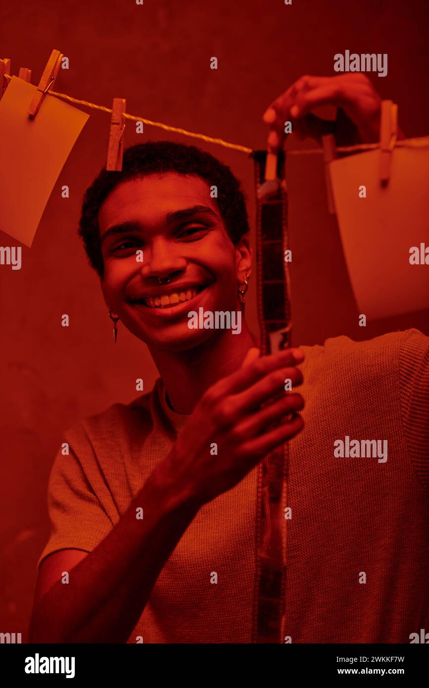 Glücklicher afroamerikanischer Mann, der frisch entwickelten Filmstreifen in einer rot beleuchteten Dunkelkammer hängt, Nostalgie Stockfoto