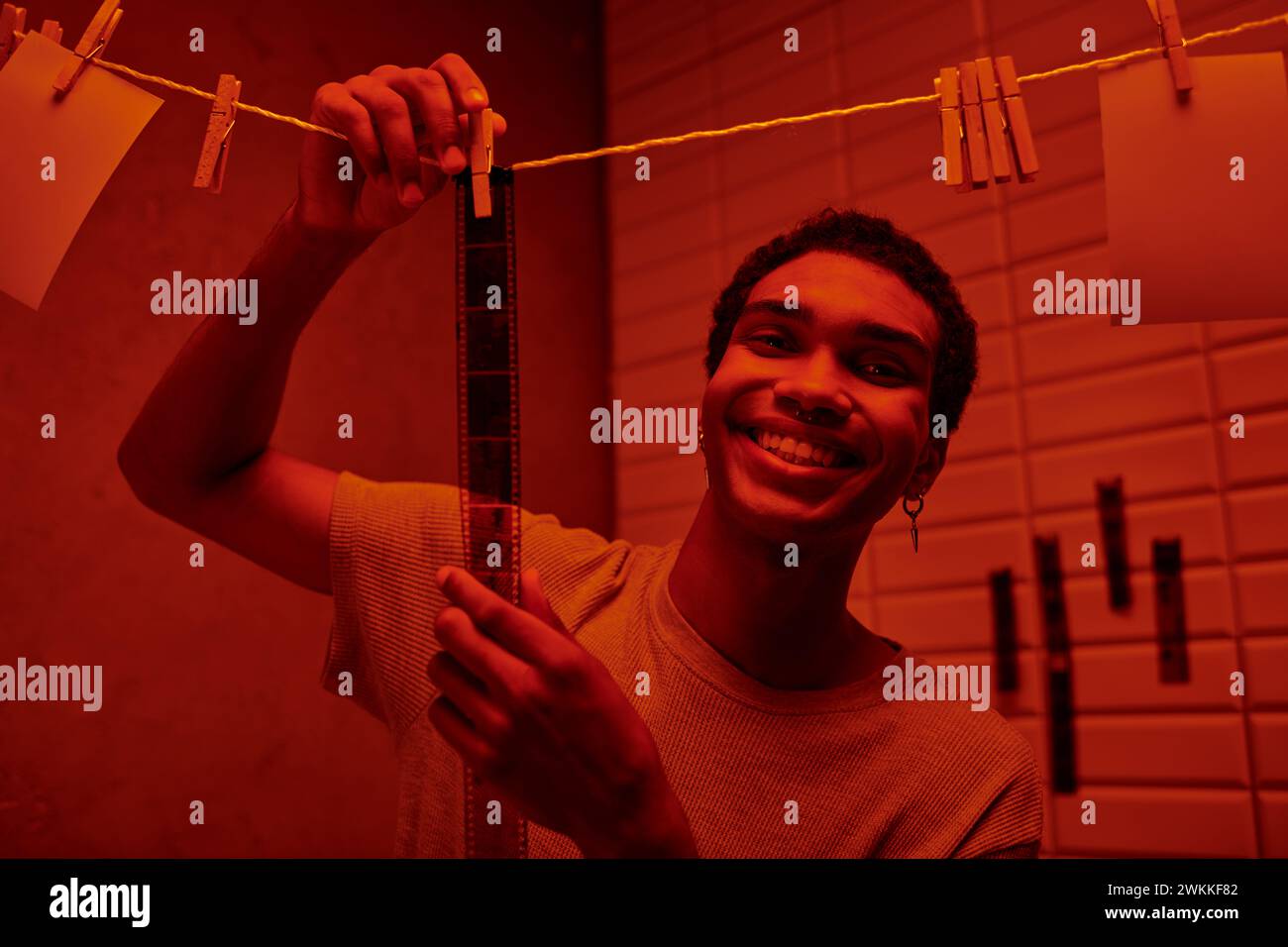 Fröhlicher afroamerikanischer Mann, der frisch entwickelten Filmstreifen in einer rot beleuchteten Dunkelkammer hängt, Nostalgie Stockfoto