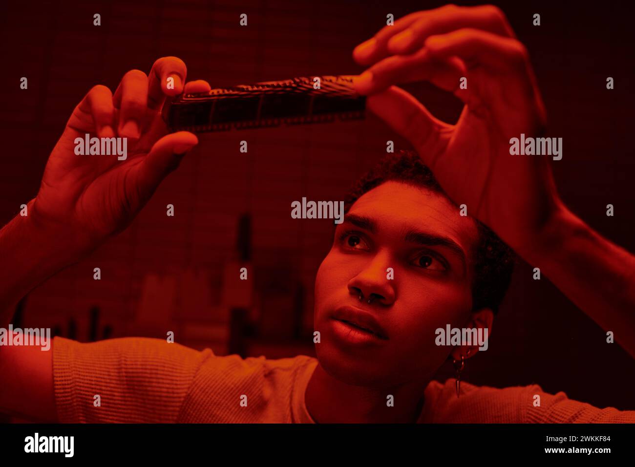 Fokussierter afroamerikanischer Fotograf, der sich einen entwickelten Filmstreifen in einer rot beleuchteten Dunkelkammer ansieht Stockfoto