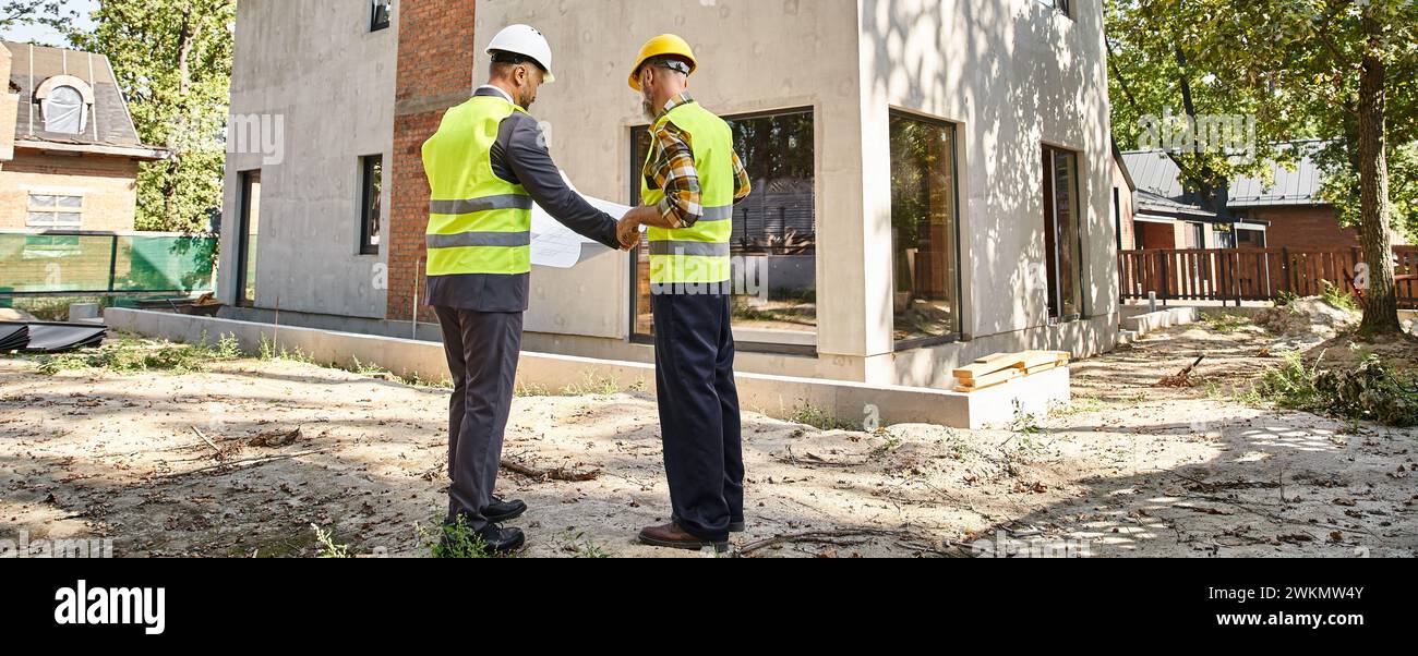 Engagierter Hüttenbauer und Architekt, der einen Plan hält und über Baustelle, Banner diskutiert Stockfoto