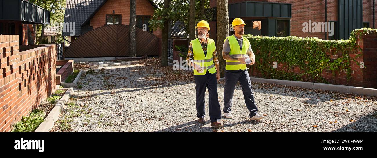 Gutaussehende Bauarbeiter in Sicherheitswesten, die auf dem Gelände spazieren gehen und diskutieren, Hüttenbauer, Banner Stockfoto