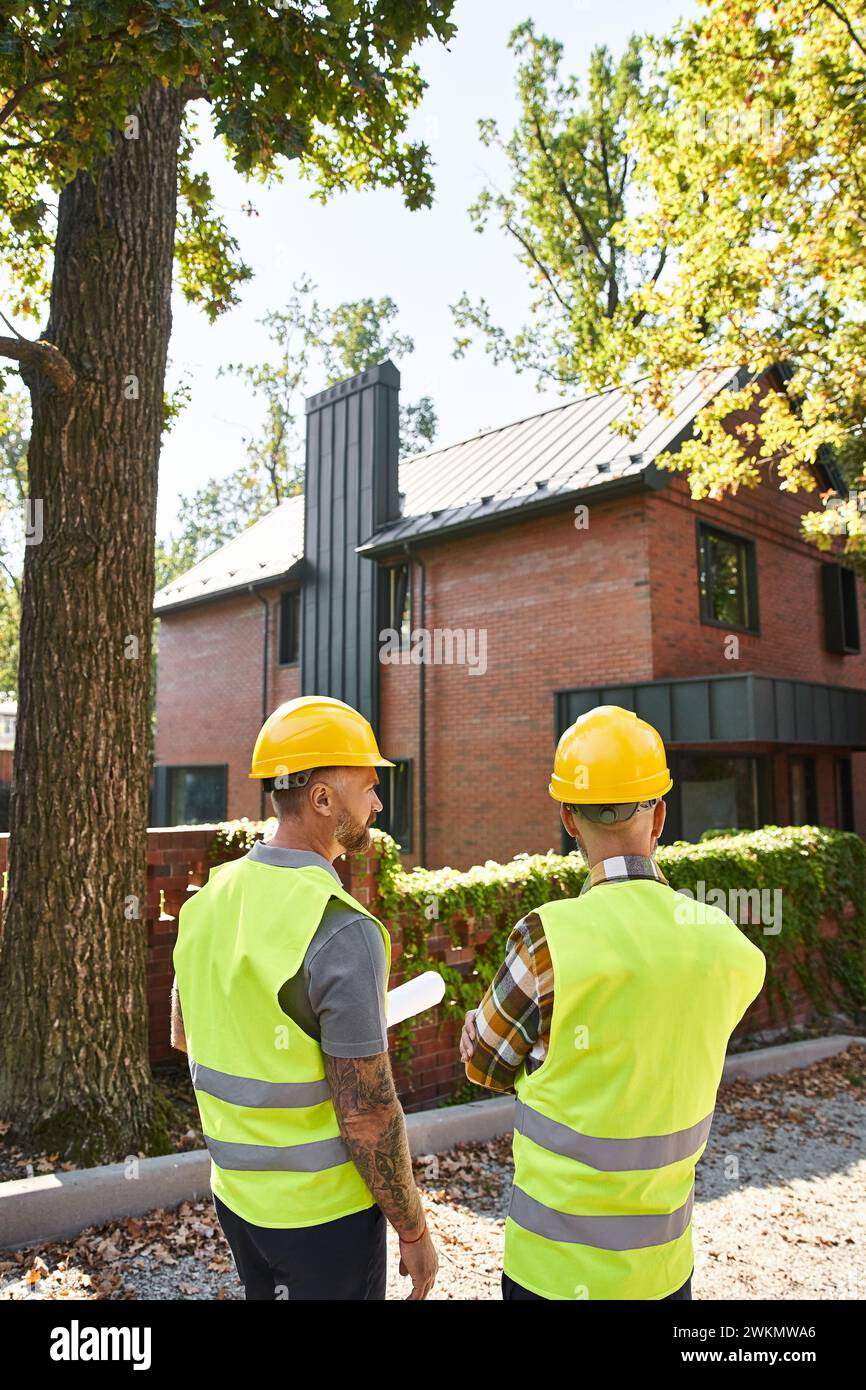 Rückansicht von engagierten Bauarbeitern in Sicherheitswesten mit Bauplan vor Ort, Hüttenbauer Stockfoto