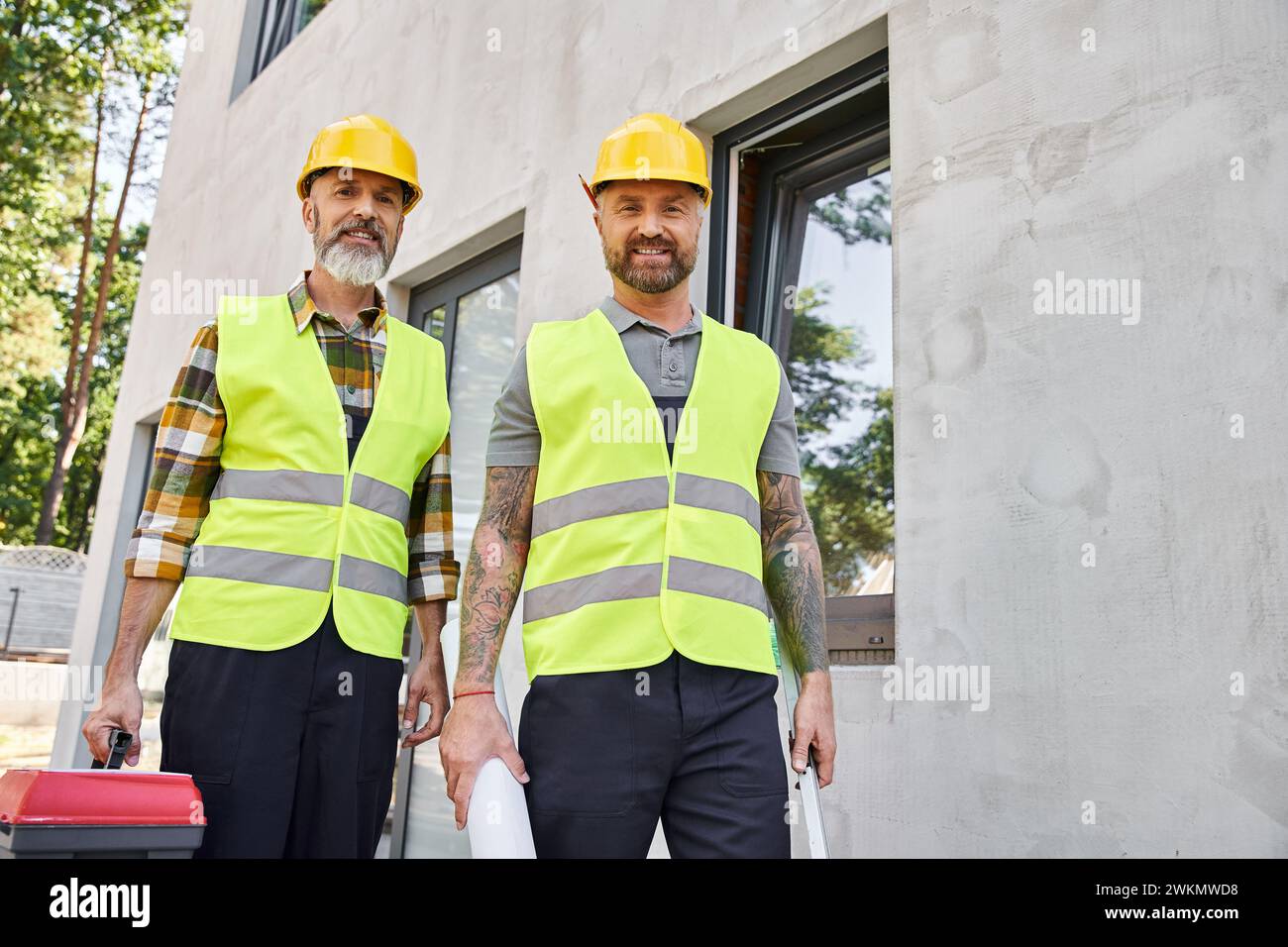 Fröhliche Bauarbeiter mit Werkzeugkastenniveau und Bauplan, die draußen in die Kamera schauen Stockfoto