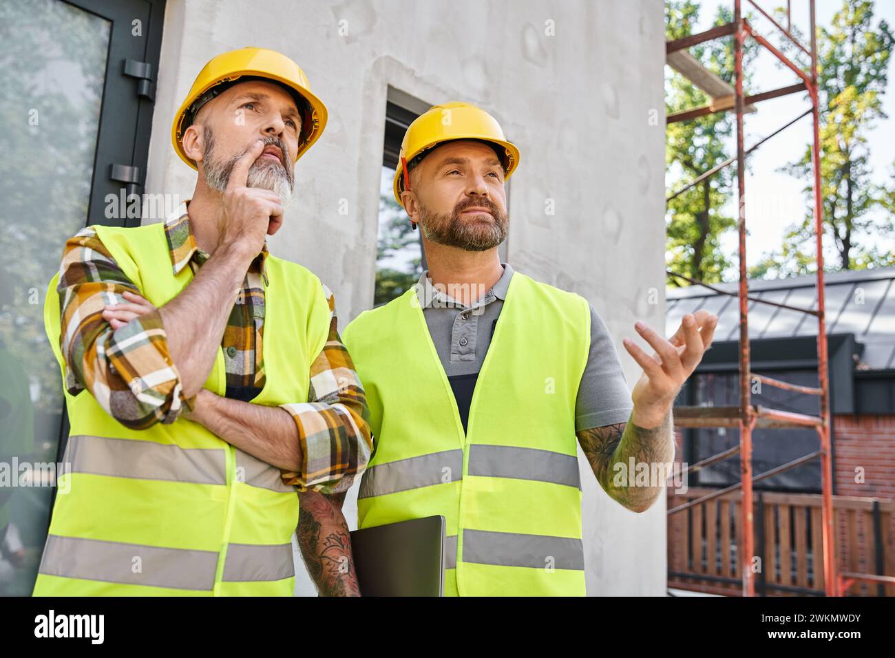 Zwei gut aussehende Bauarbeiter in Sicherheitswesten und Helmen diskutieren über die Baustelle, Hüttenbauer Stockfoto