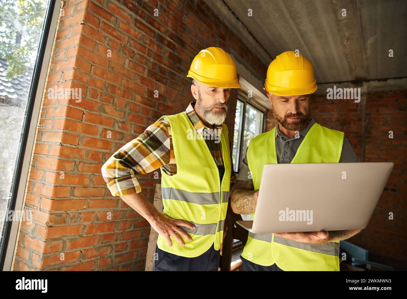 Gut aussehende Bauarbeiter in Sicherheitswesten, die vor Ort mit Laptop arbeiten, Hüttenbauer Stockfoto