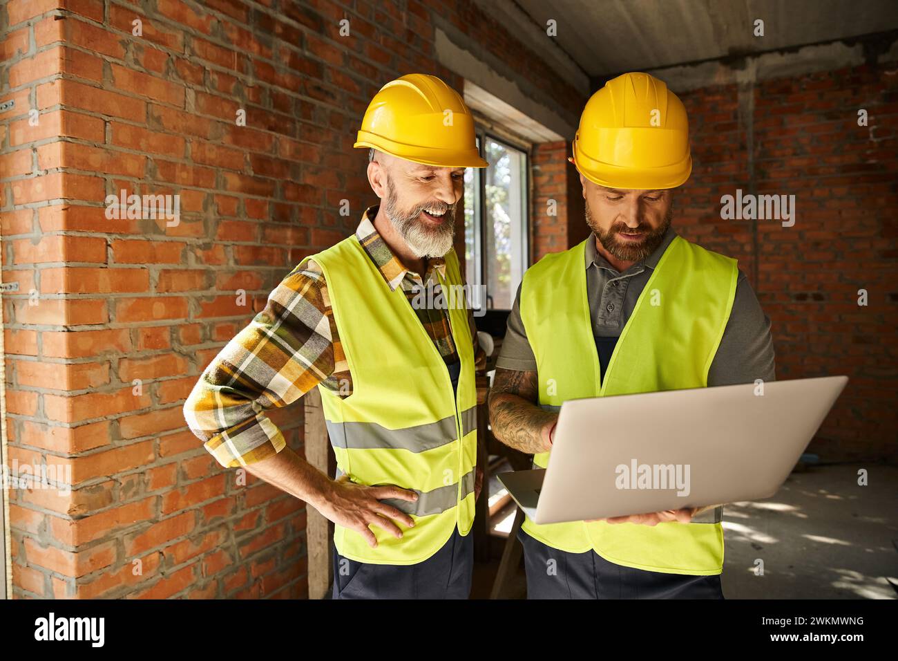 Fröhliche, hingebungsvolle Bauarbeiter in Sicherheitswesten, die vor Ort mit Laptop arbeiten, Hüttenbauer Stockfoto