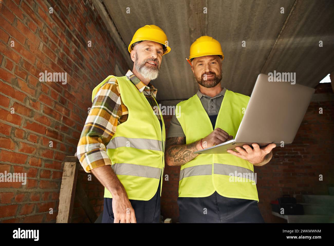 Fröhliche, hingebungsvolle Bauarbeiter in Sicherheitswesten, die vor Ort mit Laptop arbeiten, Hüttenbauer Stockfoto