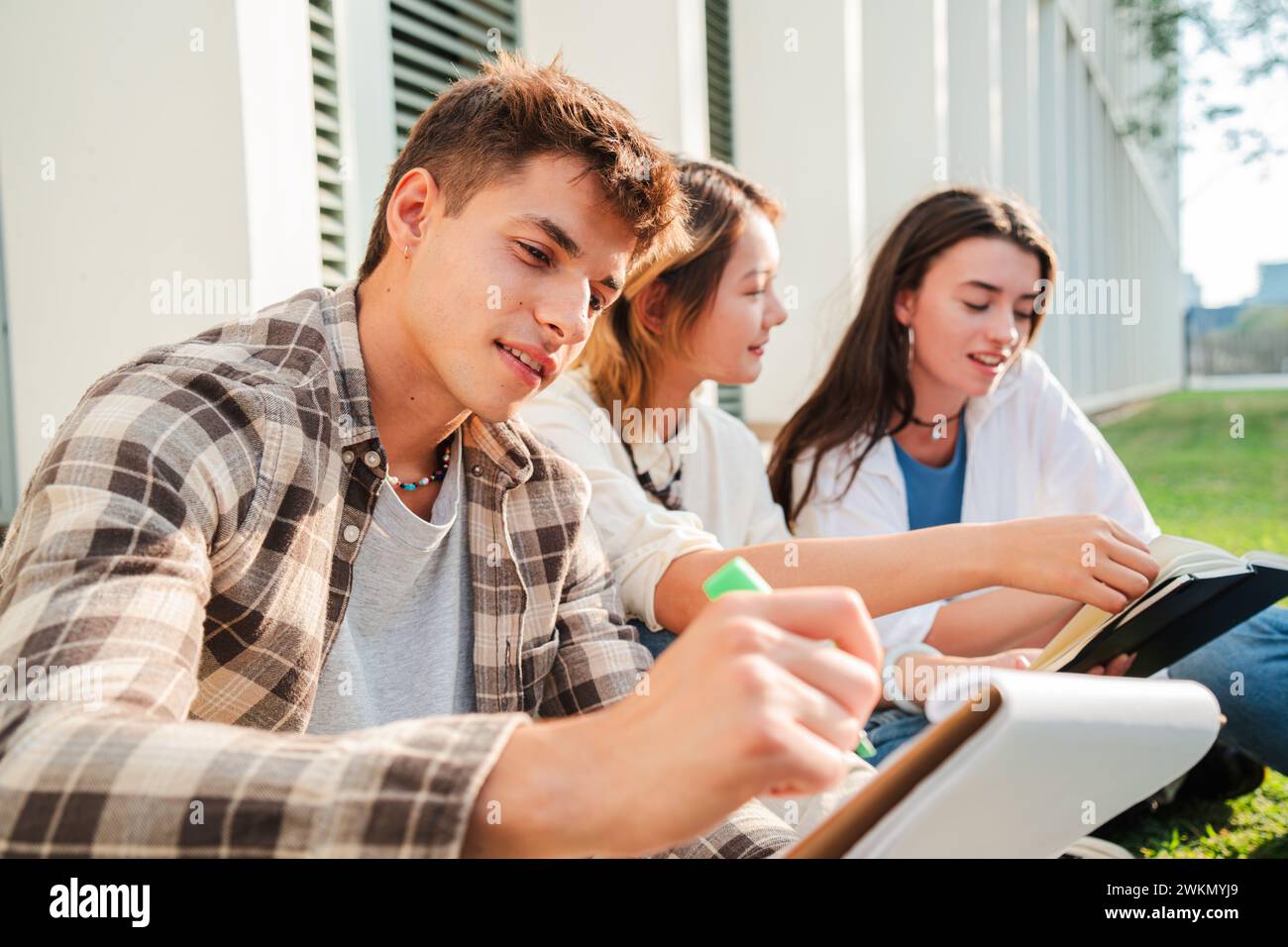 Ein junger Teenager schreibt Notizen mit einem Stift auf sein Notizbuch, sitzt mit seinen Klassenkameraden auf dem Campus-Rasen. Der High School-Typ liest und studiert die Klassenstunde für die Prüfung. Teamwork-Freunde. Hochwertige Fotos Stockfoto