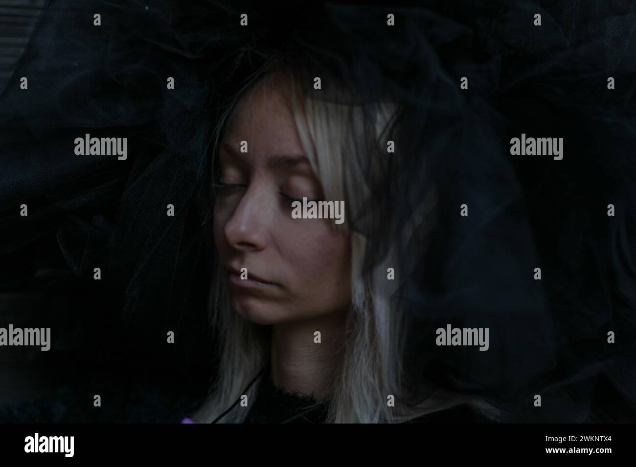 Das Gesicht einer jungen kaukasischen blonden Frau ist teilweise von einem dunklen Schleier verdeckt, ihre Augen sind in einem ruhigen Ausdruck geschlossen Stockfoto