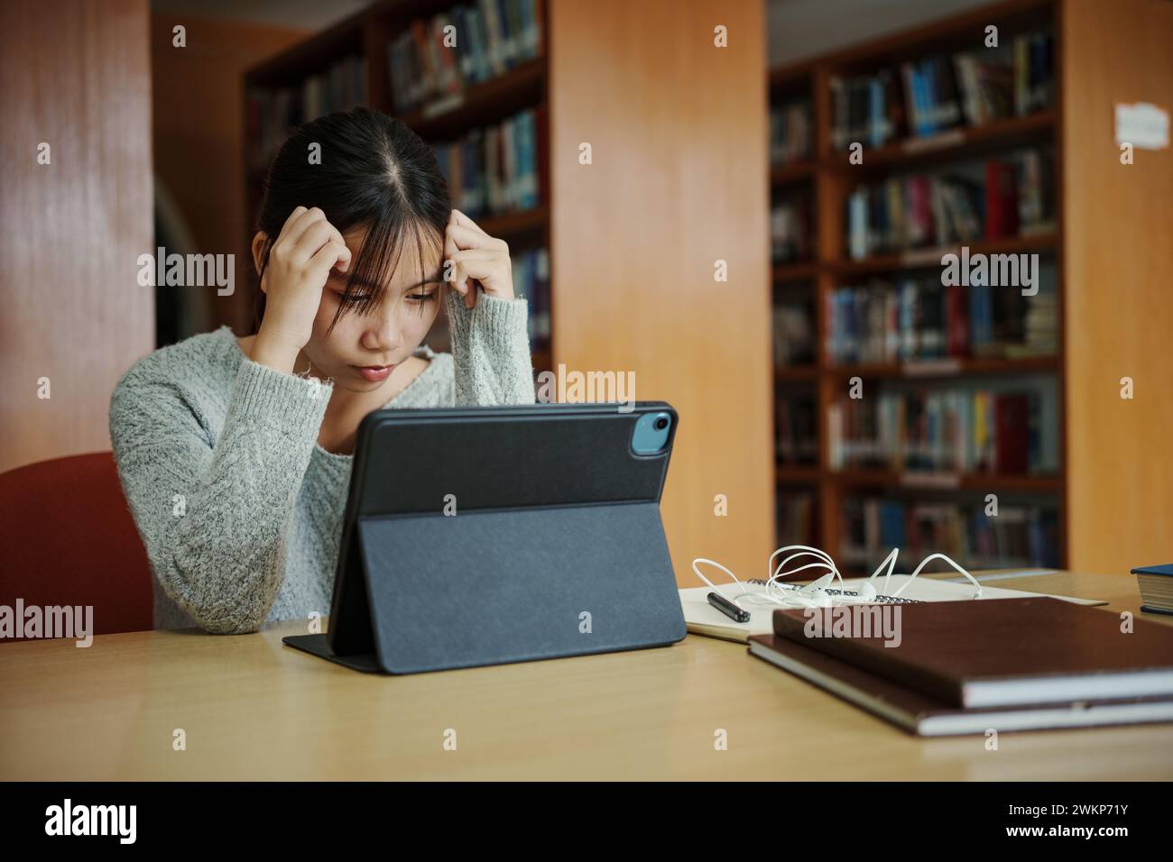 Gestresste junge Studentin sitzt am Holztisch und liest Buch während der Prüfungsvorbereitung in der Bibliothek. Stockfoto